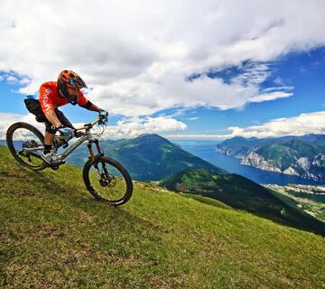 Mountain biker con casco su una mountain bike su un prato di montagna verde, con vista sul paesaggio montano circostante, sul Lago di Garda e sul cielo azzurro.