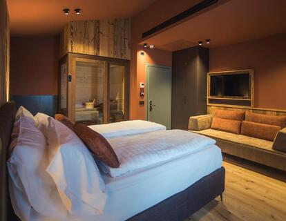 Modernes Hotelzimmer mit Doppelbett, Holzwänden, warmen Lichtakzenten, Sofa, Fernseher und Blick in die Sauna