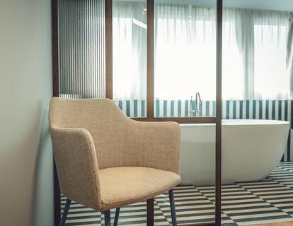 Suite Aura con poltrona imbottita beige davanti a una parete in vetro con vista sul bagno e sulla vasca freestanding.