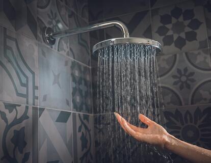 Hand unter fließendem Wasser einer Regendusche vor einer dunkel gemusterten Fliesenwand.