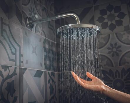 Hand unter fließendem Wasser einer Regendusche vor einer dunkel gemusterten Fliesenwand.