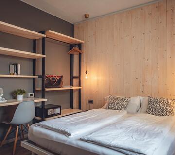 Camera d'hotel moderna con letto matrimoniale, scaffale aperto, scrivania e parete in legno illuminata da una luce calda.