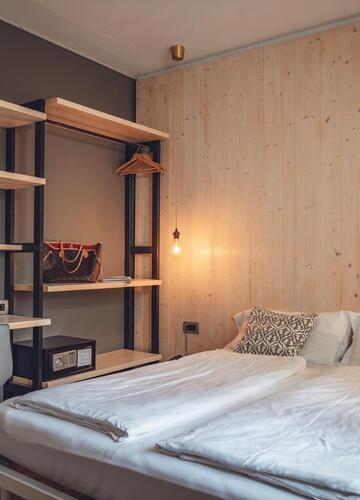 Modernes Hotelzimmer mit Doppelbett, offenem Regal, Schreibtisch und warm beleuchteter Holzwand.