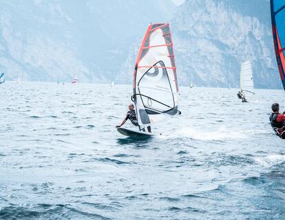 Diversi windsurfisti con vele colorate sul Lago di Garda, con il paesaggio montano circostante sullo sfondo.