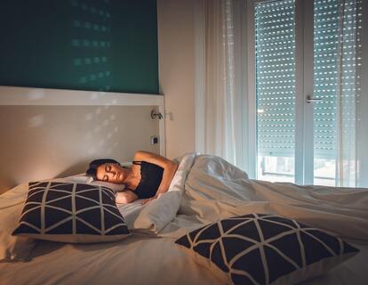 Frau liegt entspannt auf einem Doppelbett mit weißer Bettwäsche und dunkel gemusterten Kissen in einem Hotelzimmer mit Balkon.