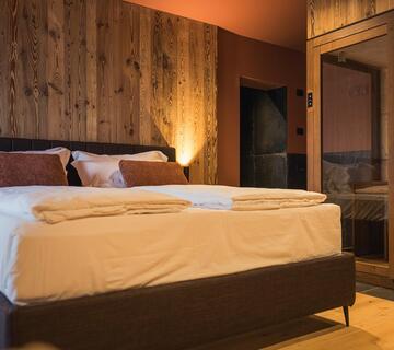 Camera moderna d’hotel con letto matrimoniale, biancheria da letto bianca, testiera imbottita, pavimento in legno, parete in legno illuminata e sauna con pareti in vetro.