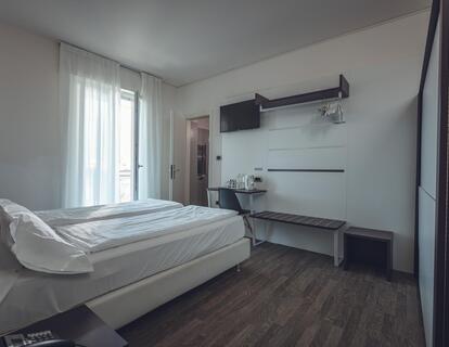 Modernes Zimmer im Hotel Villa Alberta mit Doppelbett, weißer Bettwäsche, Holzboden, Schreibtisch mit Stuhl, Wasserkocher, Fernseher, Wandgarderobe und Balkontür.