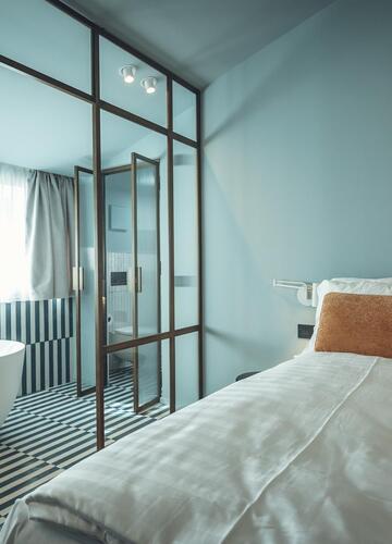 Suite Aura con letto matrimoniale, pareti azzurre, pavimento piastrellato con motivi in bianco e nero e vista sulla vasca da bagno freestanding dietro una parete in vetro.