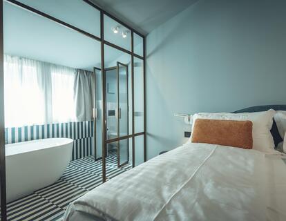 Suite Aura mit Doppelbett, hellblauen Wänden, schwarz-weiß gemustertem Fliesenboden und Blick auf die freistehende Badewanne hinter einer Glaswand.