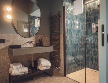Modern bathroom at Hotel Villa Alberta with washbasin, large wall mirror and shower with dark blue tiles.