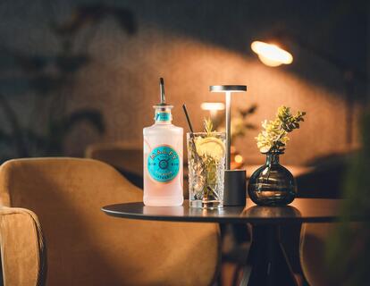 Accogliente zona relax con poltrona imbottita, tavolo scuro, bevande e vaso di fiori in un ambiente illuminato da una luce calda.