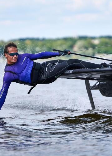 Sailor in a blue wetsuit leaning out of a sailboat and touching the surface of Lake Garda with one hand.