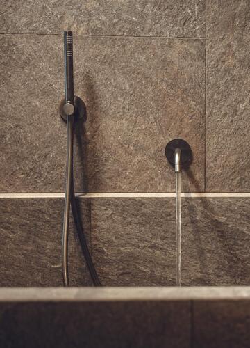 Detailaufnahme einer modernen Badewanne mit dunkel gefliesten Wänden, Wandarmatur und Handbrause.