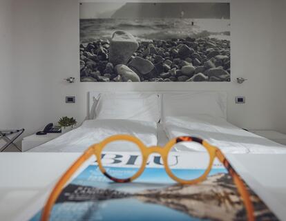Helles Hotelzimmer mit Doppelbett, großem Schwarz-Weiß-Bild und einer Brille auf einem Buch im Vordergrund.