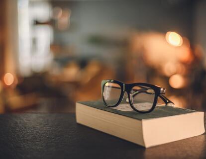 Occhiali da lettura neri su un libro chiuso, appoggiato su un tavolo scuro.