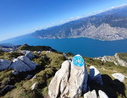 Rocky viewpoint with a trail marker and panoramic views of Lake Garda and the surrounding mountain landscape.