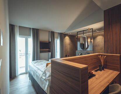 Suite Atlas with double bed, wooden furniture, warm lighting accents, television and large window with curtains.