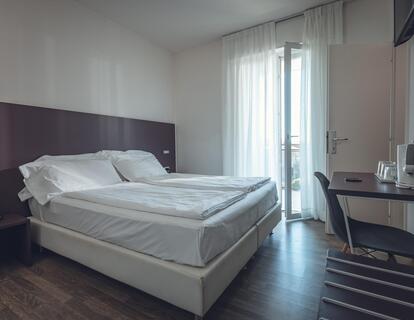 Modernes Zimmer im Hotel Villa Alberta mit Doppelbett, weißer Bettwäsche, Holzboden, Schreibtisch mit Stuhl, Wasserkocher, Fernseher und großer Balkontür.