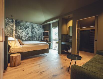 Suite Dryas with wooden flooring, a double bed, floral wallpaper, a seating area with a sofa and a view of the bathroom area.
