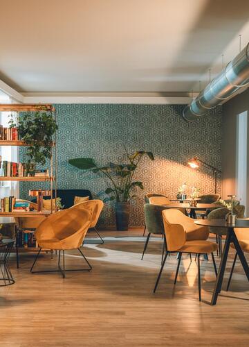 Accogliente sala comune con poltrone, tavoli, libreria e piante in un ambiente luminoso e arredato in stile moderno.