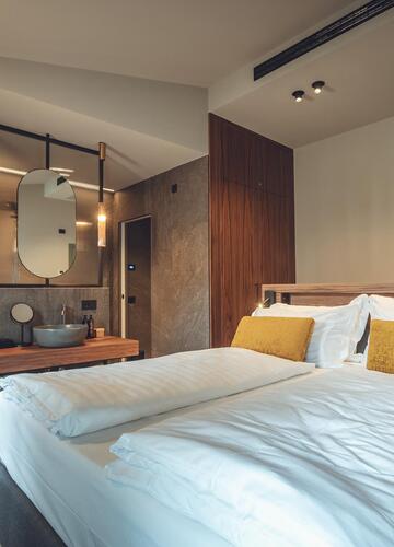 Atlas Suite with double bed, warm lighting accents and a view into the adjoining bathroom with washbasin, round mirror and bathtub.